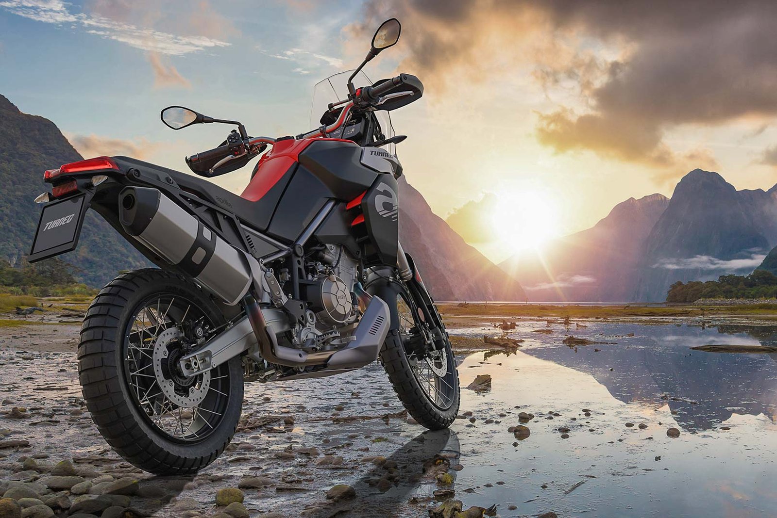 Moto Aprilia tuareg 660 rossa e nera senza targa ferma ai bordi di un fiordo con il sole al tramonto tra le montagne in lontananza